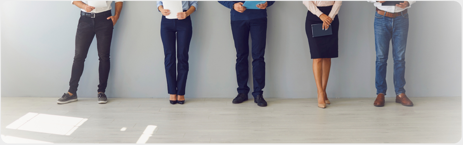 書類を手に持っている5人の人物が壁の前で並んで立っている